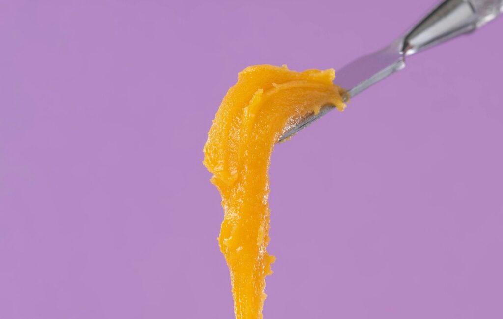 Creamy orange badder cannabis concentrate dripping from a metal dab tool on a purple background