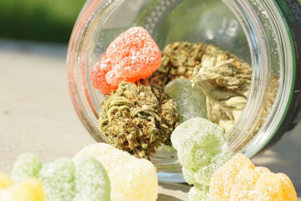 Sugar-coated cannabis gummies spilling from a glass jar with cannabis flower buds