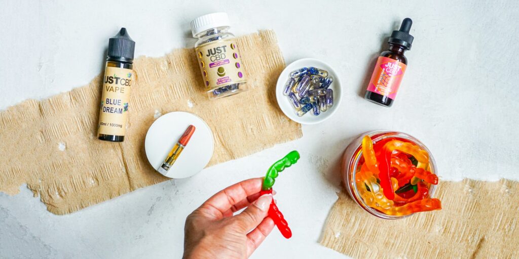 Hand holding a gummy worm above a jar of gummies, next to a tincture bottle, capsules and a vape cartridge