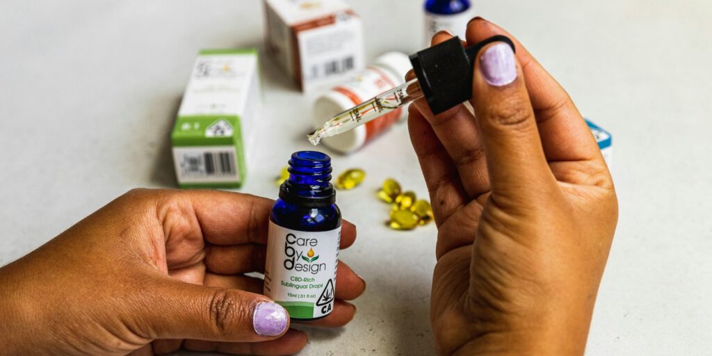 Hands holding a sublingual cannabis tincture bottle and a filled glass dropper, with softgel capsules behind
