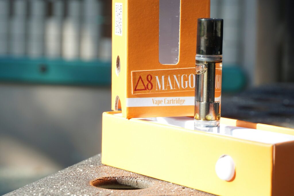 Glass vape cartridge filled with golden cannabis oil standing in its open orange box